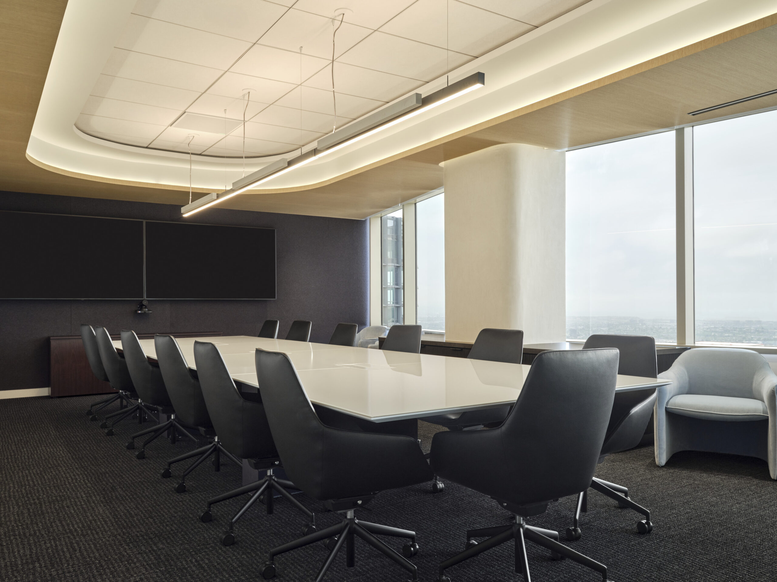 A sleek, modern boardroom featuring a long, white conference table surrounded by ergonomic black chairs. Soft, curved LED lighting enhances the ceiling's architectural lines, while large windows offer panoramic city views, creating an inviting and professional atmosphere for meetings and collaborations. Dark gray walls and carpet provide contrast.