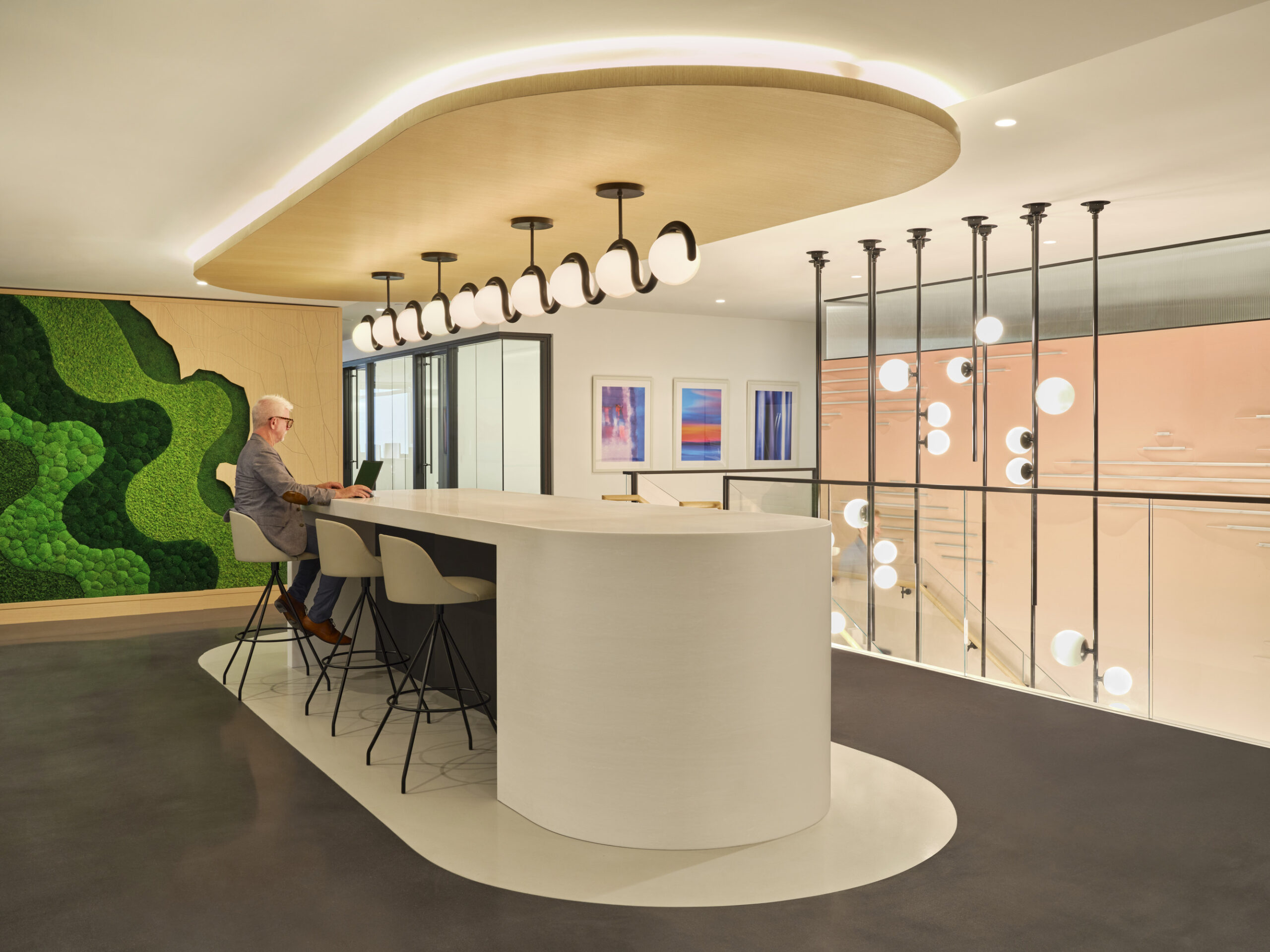 A modern workspace featuring a striking curved white countertop and minimalist bar stools. Above, contemporary circular pendant lights create a warm atmosphere. A vibrant green moss wall adds a pop of color, complementing the neutral tones of the floor and walls, enhancing the inviting and innovative design.