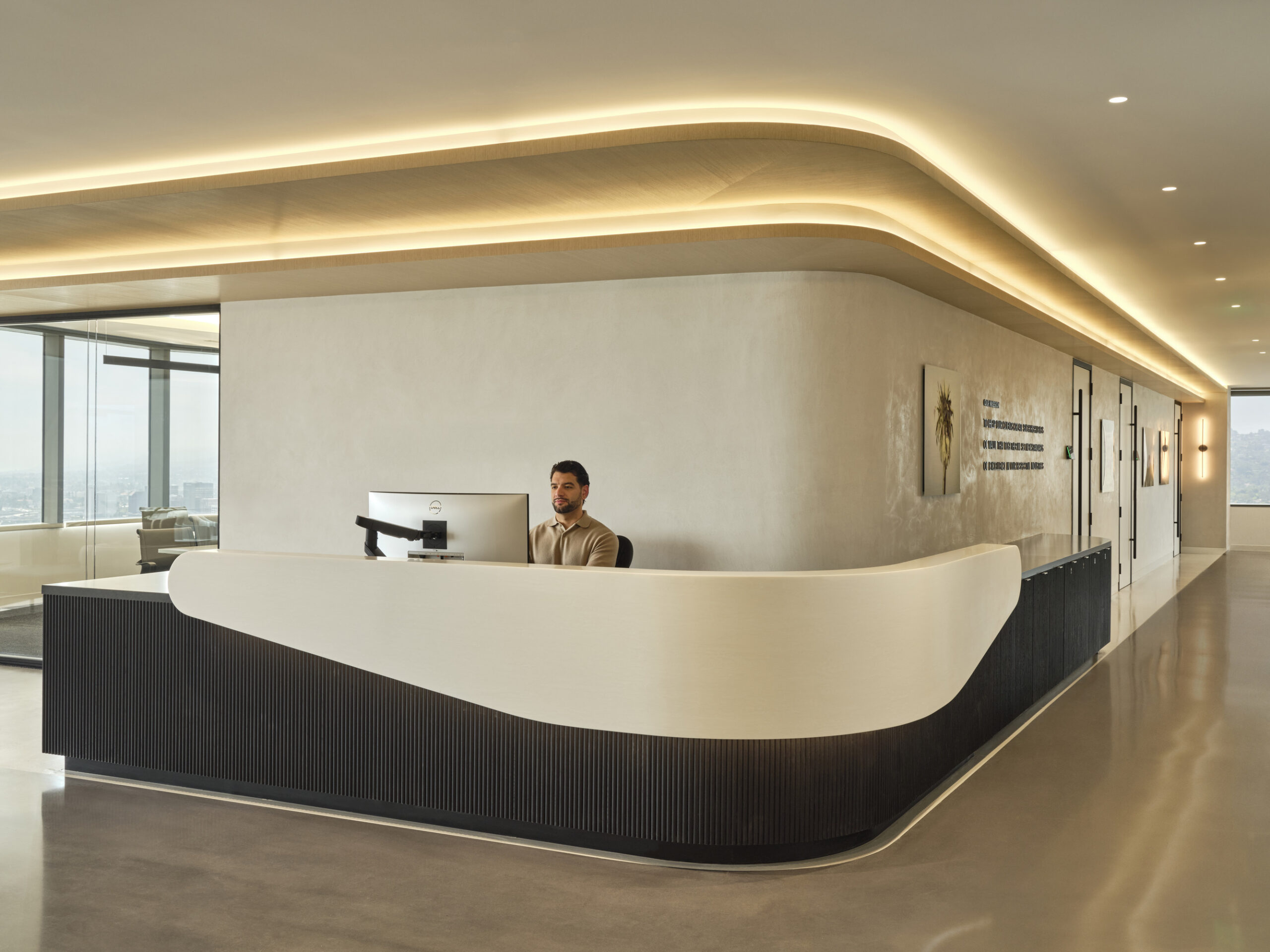 The reception area features a sleek, curved desk with a smooth, sculptural quality, contrasting with a textured black base. Soft, ambient lighting highlights the minimalist design while large windows offer views of a city skyline, creating an inviting and sophisticated atmosphere in a modern office space.