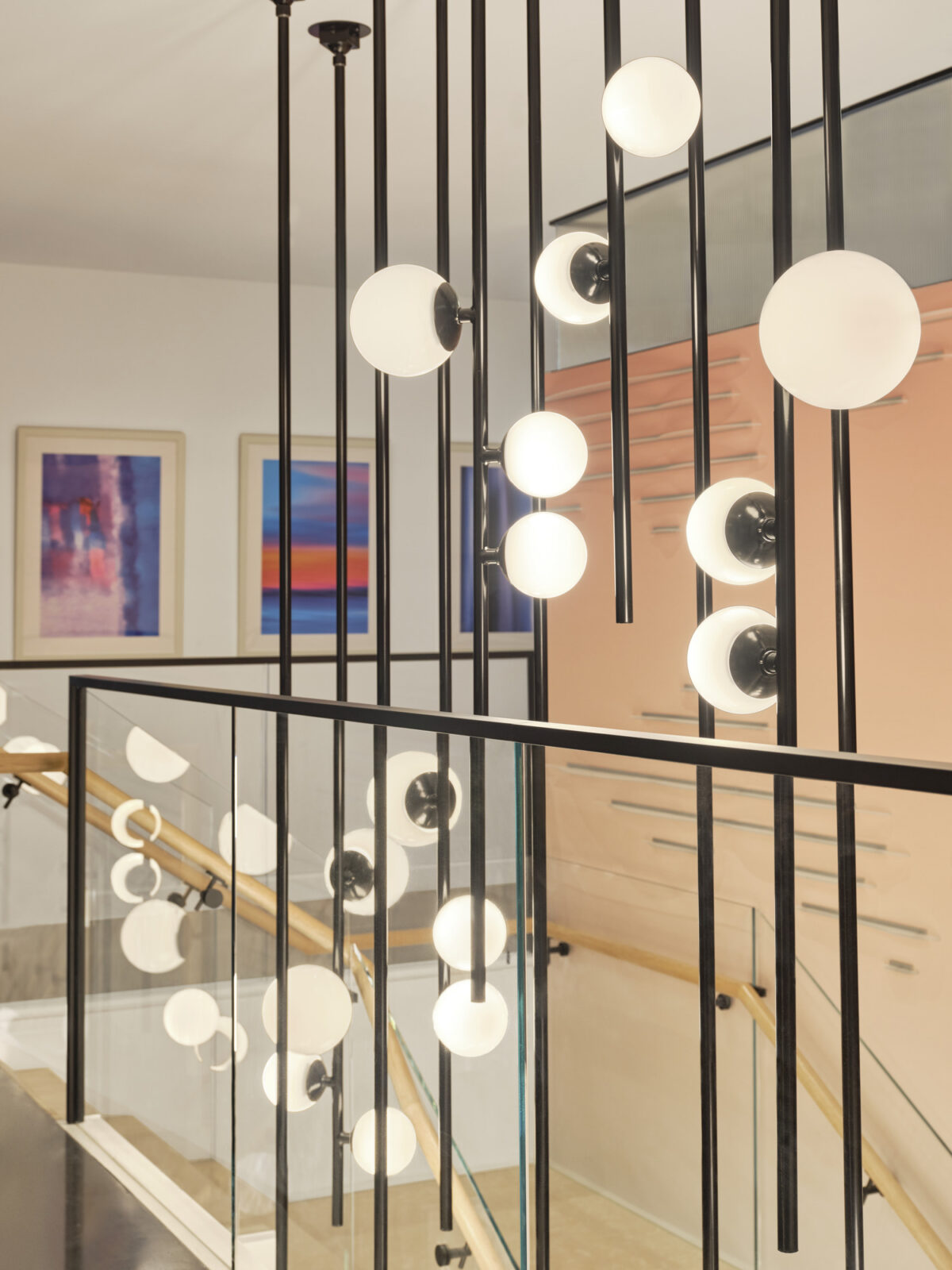 Suspended black metal lighting fixtures with spherical white glass globes create a striking visual centerpiece above a modern staircase. The elegant design contrasts with the minimalist railing and colorful framed artwork on the walls, enhancing the contemporary ambiance of the interior space.