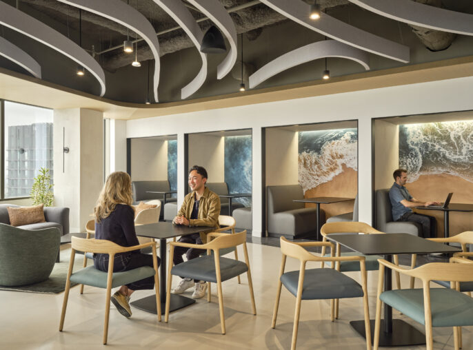 A modern, open-concept lounge area features a mix of seating options including sleek wooden chairs and plush, rounded sofas. Waves of sculptural ceiling elements mimic ocean waves, while large murals depicting seascapes enhance the calming atmosphere. Natural light floods the space, promoting an inviting, collaborative environment.