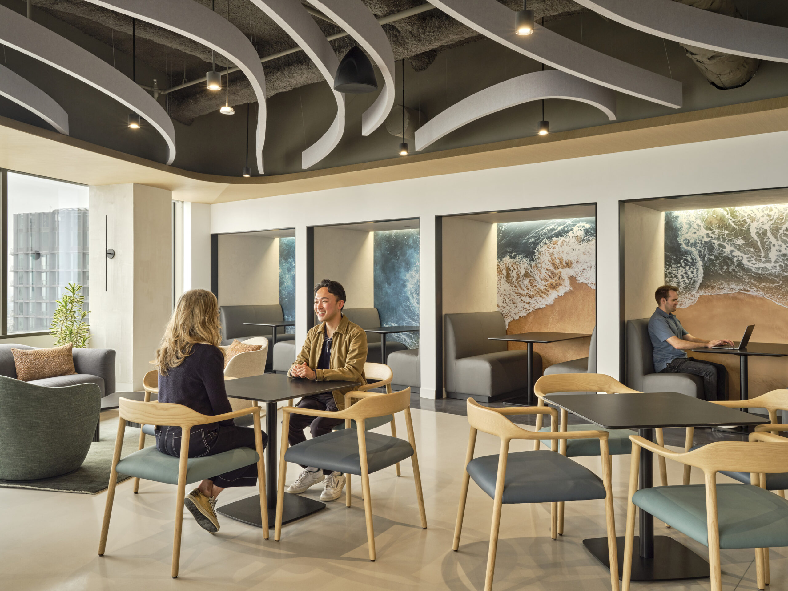 A modern, open-concept lounge area features a mix of seating options including sleek wooden chairs and plush, rounded sofas. Waves of sculptural ceiling elements mimic ocean waves, while large murals depicting seascapes enhance the calming atmosphere. Natural light floods the space, promoting an inviting, collaborative environment.
