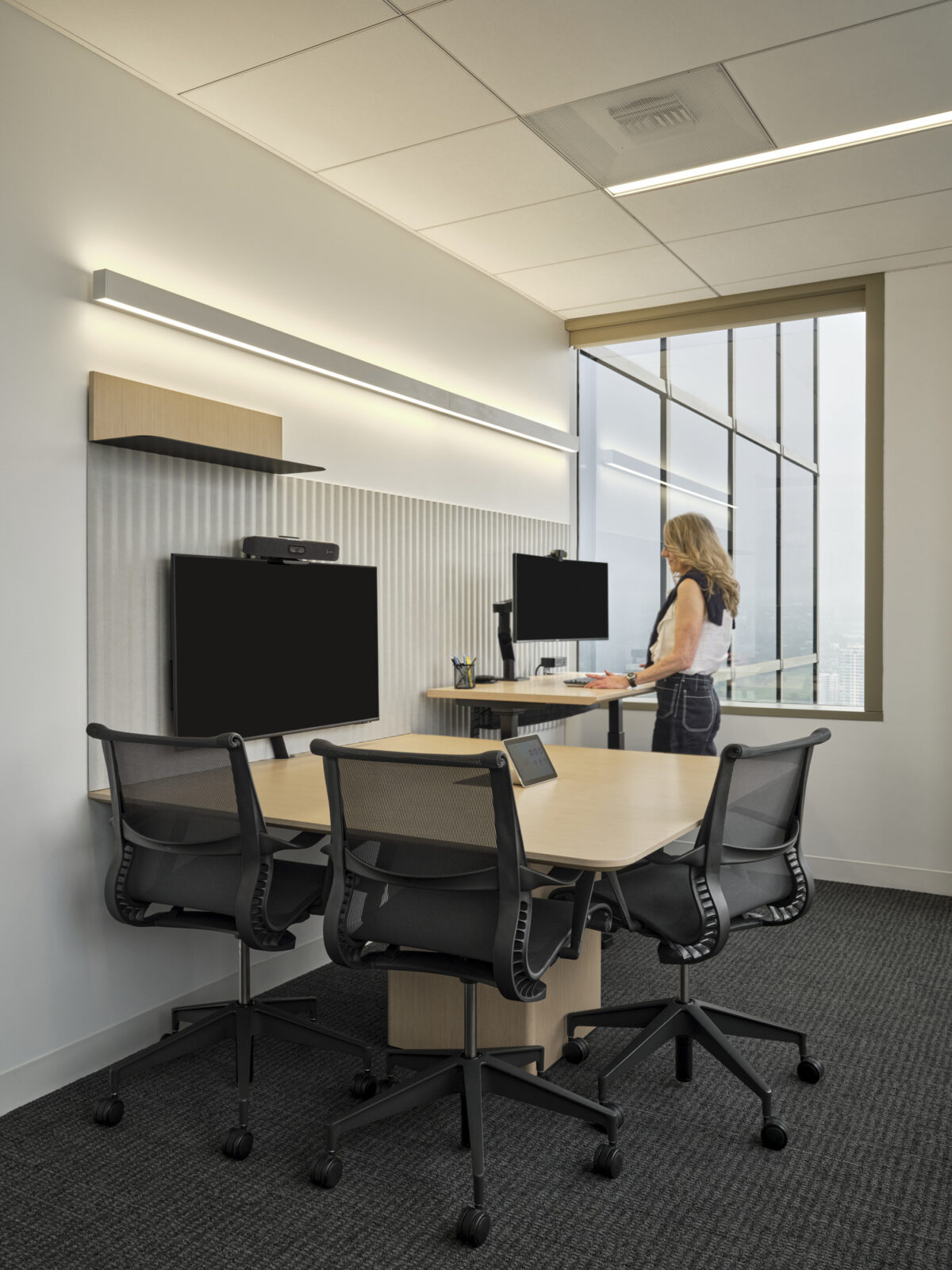 This workspace features a sleek, modern design with minimalist furniture. A light wood table is complemented by ergonomic black chairs, while integrated technology, including dual monitors and a video conferencing unit, enhances collaboration. Natural light floods the room through large windows, offering a stunning cityscape view.