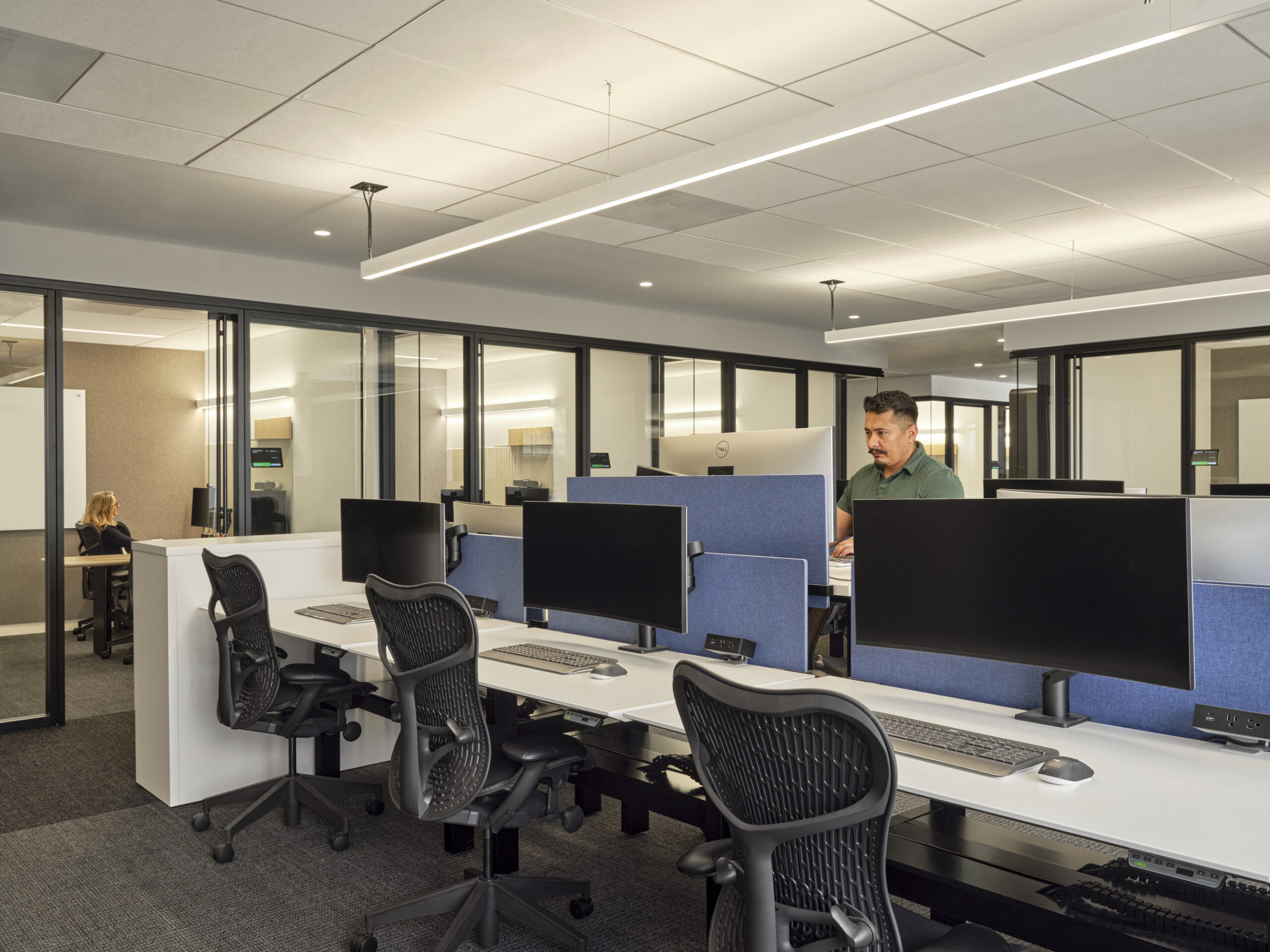 Contemporary open office space features sleek white workstations equipped with dual monitors and ergonomic chairs. Glass partitions enhance natural light and promote collaboration while maintaining privacy. Neutral tones and modern lighting create a professional atmosphere conducive to productivity and creativity.