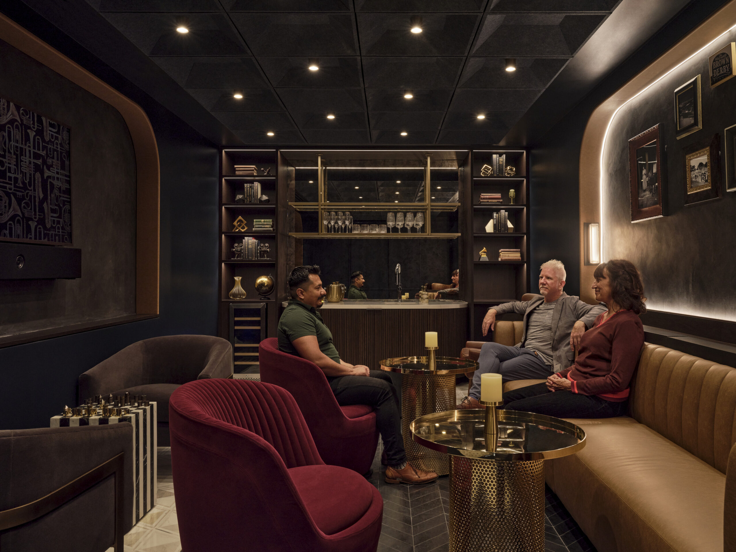 A stylish lounge features a rich color palette of deep blues and gold accents. Plush velvet seating in burgundy and beige complements the geometric ceiling design. Elegant artwork adorns the walls, while a softly lit bar area invites conversation, creating an intimate yet sophisticated atmosphere ideal for social gatherings.