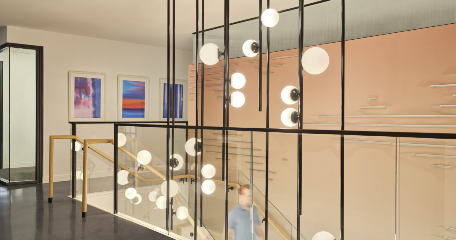A modern staircase features sleek glass railings and dramatic pendant lighting with minimalist round bulbs, suspended from black poles. Subtle artwork adorns the walls, creating a sophisticated atmosphere. The overall design emphasizes open space and contemporary aesthetics, enhancing the flow of natural light throughout the area.
