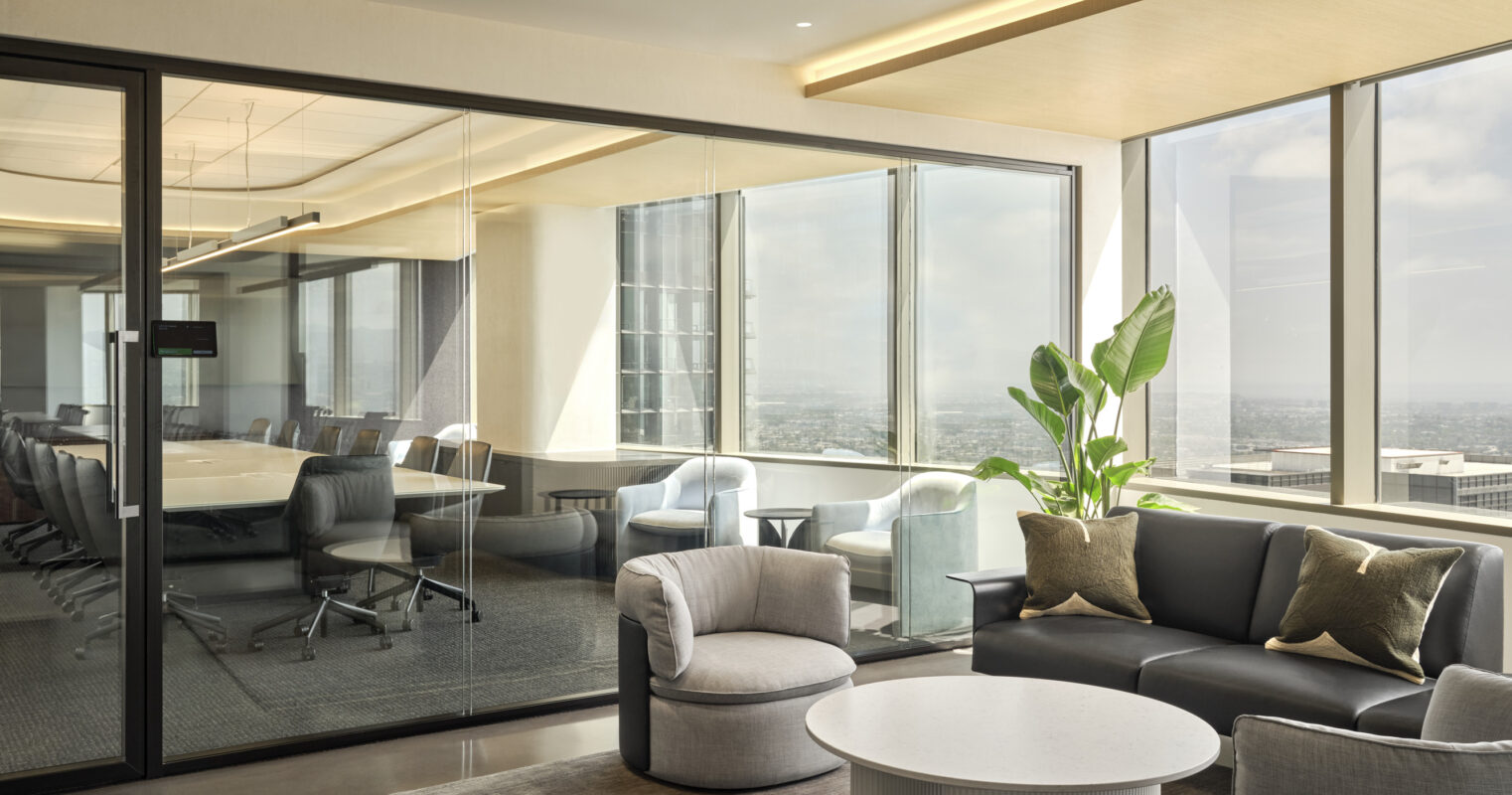A modern office lounge featuring a sleek black sectional sofa adorned with decorative cushions. A round, white coffee table sits atop a textured rug, complemented by a curved armchair. Floor-to-ceiling glass walls offer a panoramic view, while greenery adds vitality to the sophisticated atmosphere.