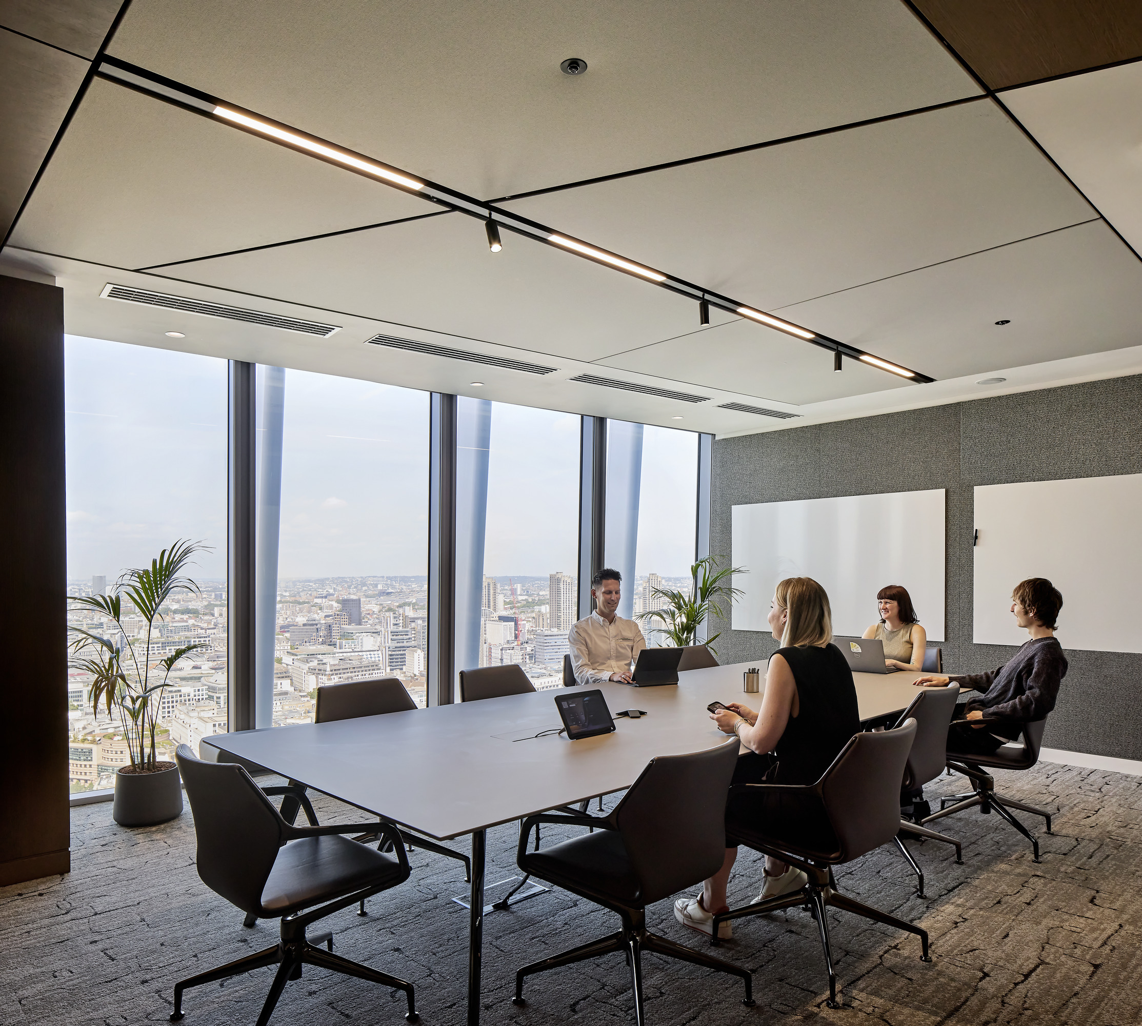 A modern conference room features a sleek, elongated table surrounded by ergonomic chairs. Large floor-to-ceiling windows offer panoramic city views, while recessed lighting enhances the clean, minimalist aesthetic. The walls combine textured panels and whiteboards, fostering a collaborative and dynamic atmosphere for meetings.