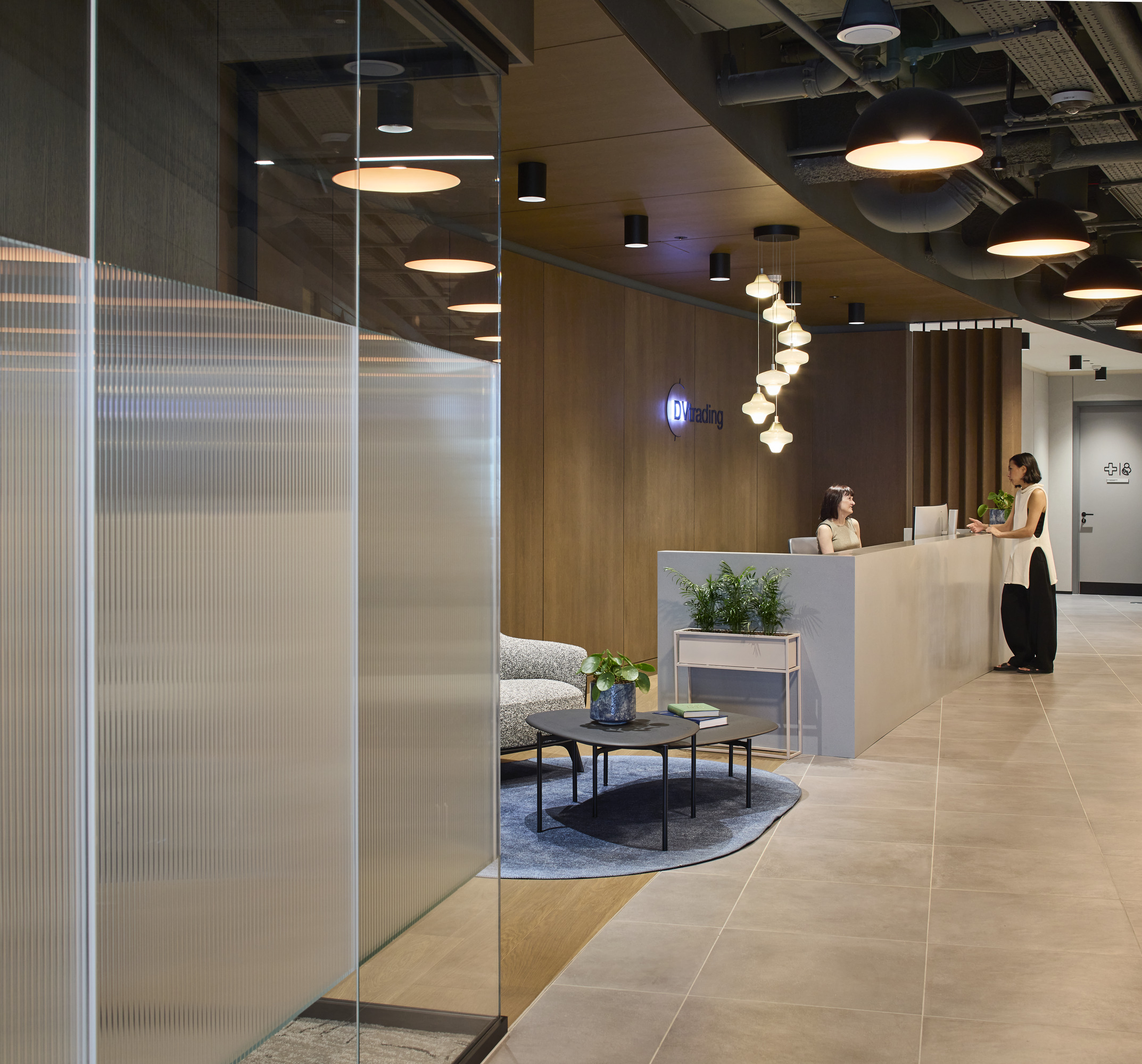 Modern reception area featuring sleek wood paneling and minimalist design. A reception desk offers a warm welcome, complemented by stylish seating and greenery. Unique pendant lighting creates an inviting atmosphere, while large windows allow natural light to enhance the space, promoting a sense of openness and professionalism.