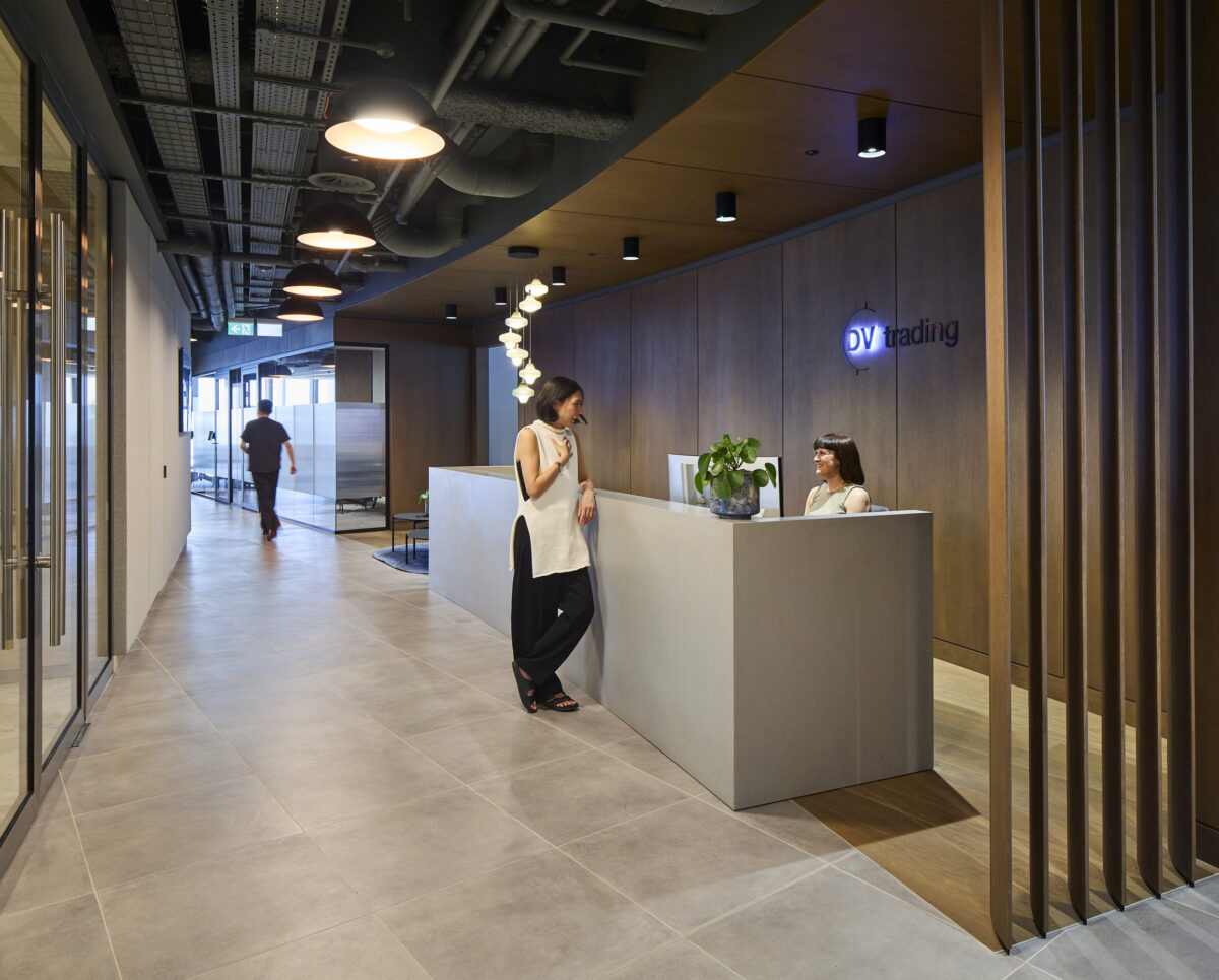 The reception area features a minimalist design with a large, smooth reception desk in light gray, complemented by warm wood accents on the walls. Natural light streams through glass partitions, adding a contemporary touch. Two staff members engage casually while a visitor walks through the space, showcasing a welcoming atmosphere.