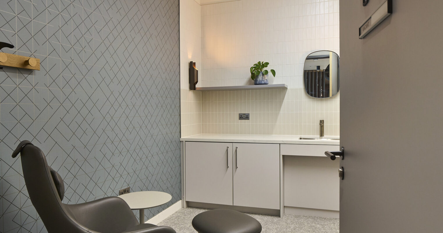 Stylish private waiting area featuring a modern gray accent wall with a geometric pattern. A contemporary black armchair complements a round ottoman, while a sleek white kitchenette with a circular mirror adds functionality. Soft overhead lighting enhances the inviting ambiance, perfect for a calming environment.