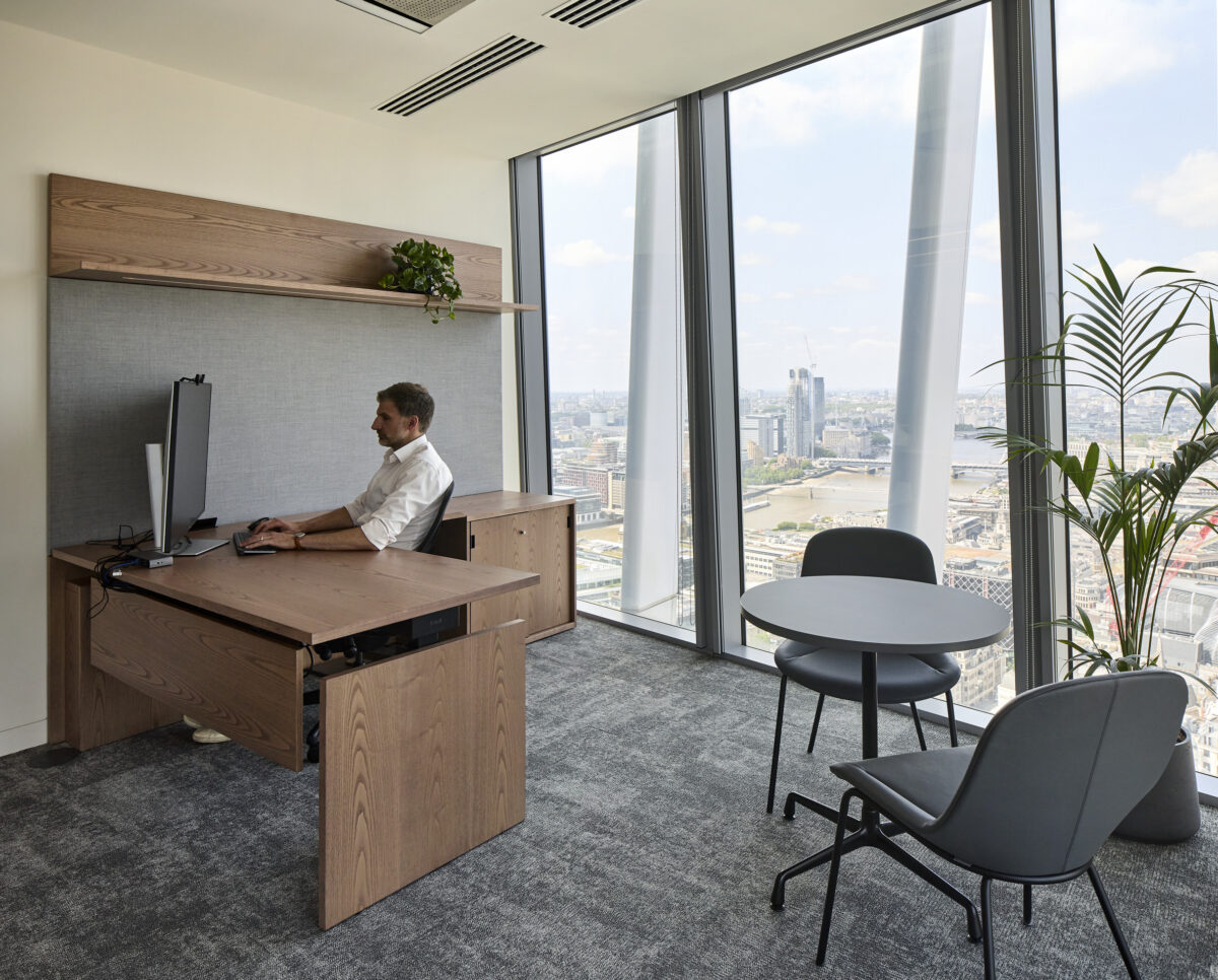 Modern office space featuring a sleek wooden desk with integrated storage, paired with a minimalist round table and contemporary chairs. A large window showcases a panoramic city view, enhancing the workspace's natural light. Greenery adds a touch of warmth, promoting a productive and inviting atmosphere.