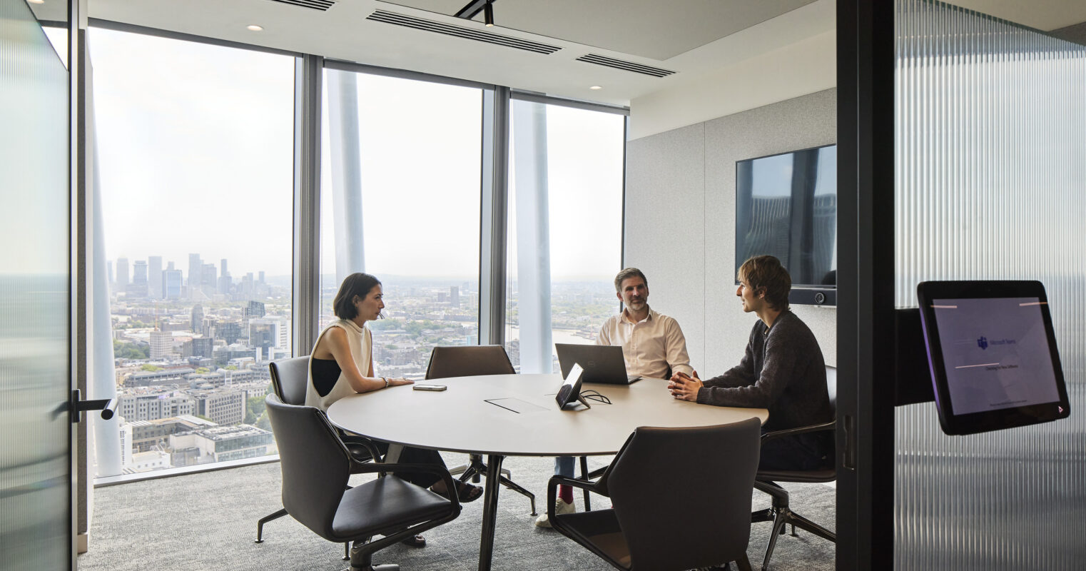 A modern conference room featuring a large circular table surrounded by ergonomic chairs. Three professionals engage in discussion, while floor-to-ceiling windows provide a panoramic view of the city skyline. The minimalist design focuses on functionality, enhanced by the soft, neutral color palette and contemporary furnishings.