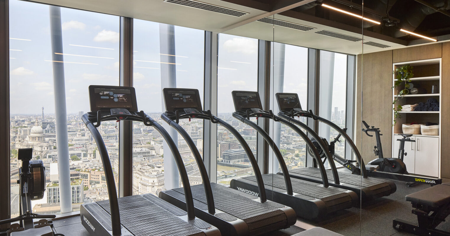 A modern gym with floor-to-ceiling windows showcasing city views. The space features sleek, black treadmills aligned against a mirrored wall. Beneath, a cushioned bench complements a row of fitness equipment, creating a stylish and functional workout environment enriched by natural light and urban scenery.