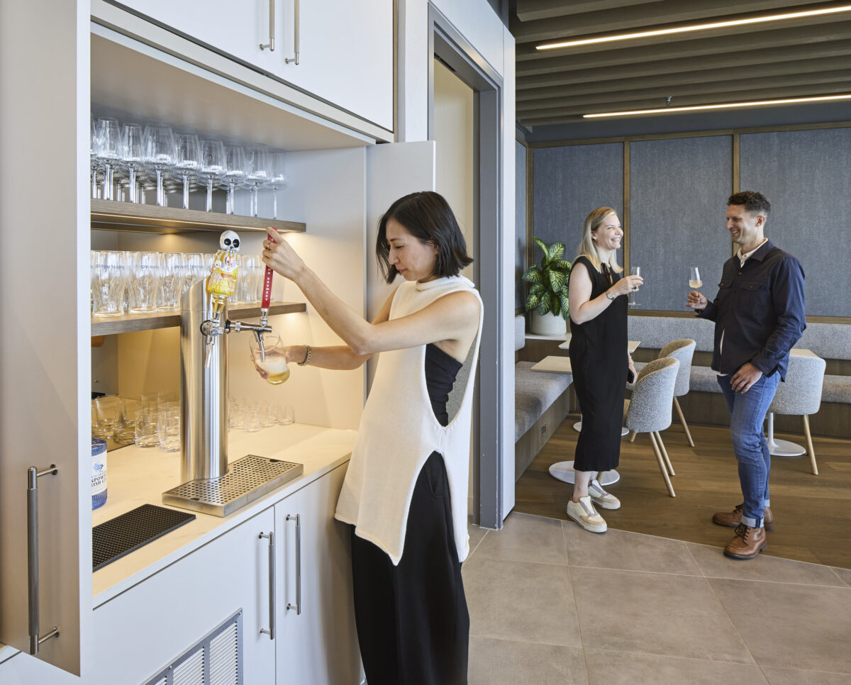 A modern bar area features a built-in tap system where a woman pours a drink, showcasing sleek cabinetry and organized glassware. In the background, two individuals engage in conversation, highlighting a warm, inviting atmosphere complemented by contemporary furnishings and tasteful decor in a spacious, well-lit room.