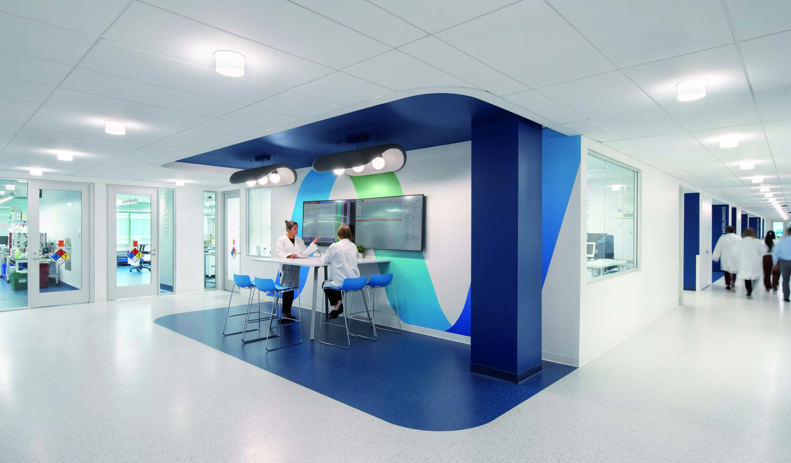 Contemporary healthcare workspace featuring a spacious hallway designed for efficient collaboration. The area showcases a striking blue and green accent wall, complemented by modern circular lighting. Ergonomic seating around a digital display fosters a productive environment for healthcare professionals, enhancing communication and workflow in a clinical setting.