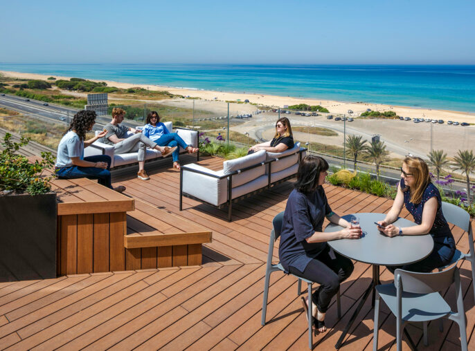 A spacious rooftop terrace featuring modern outdoor furniture, including a sectional sofa and round table. Six people are engaged in conversation, with a panoramic view of the beach and ocean. Lush greenery and wooden decking enhance the inviting atmosphere, creating a relaxing outdoor social space.