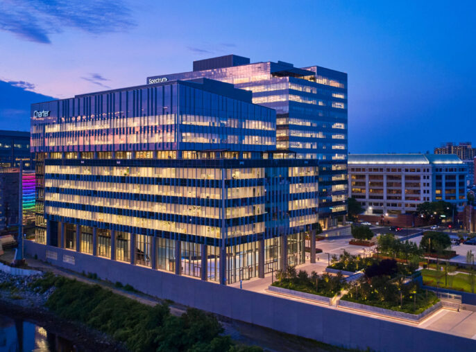 Contemporary office building featuring a striking glass façade that reflects the evening skyline. The sleek design incorporates sustainable materials and expansive windows, maximizing natural light. Surrounding landscaped areas enhance its modern aesthetic, creating an inviting atmosphere for both occupants and visitors in a vibrant urban setting.