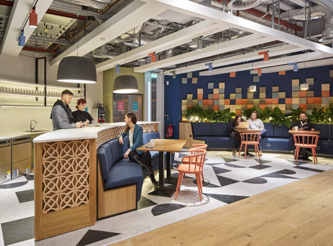 A contemporary coworking space featuring a mix of vibrant colors and textures. The communal area includes geometric black-and-white flooring, wooden accents, and patterned walls. Comfortable seating areas and a modern kitchenette enhance the space's functionality, inviting collaboration and relaxation among professionals. Lush greenery adds a refreshing touch.