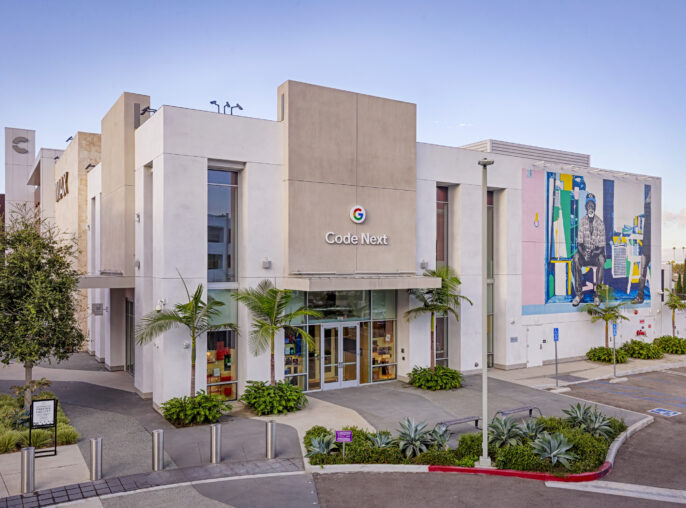 The modern facade of the Code Next building features sleek white stucco walls and expansive glass windows. A vibrant mural adorns one side, showcasing colorful figures that enhance the contemporary design. Lush landscaping, including palm trees and succulents, complements the urban environment, creating an inviting atmosphere.