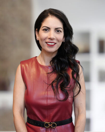 Professional portrait of a woman with long, wavy dark hair, wearing a sleeveless burgundy leather dress with a black belt, smiling against a softly blurred office interior background.
