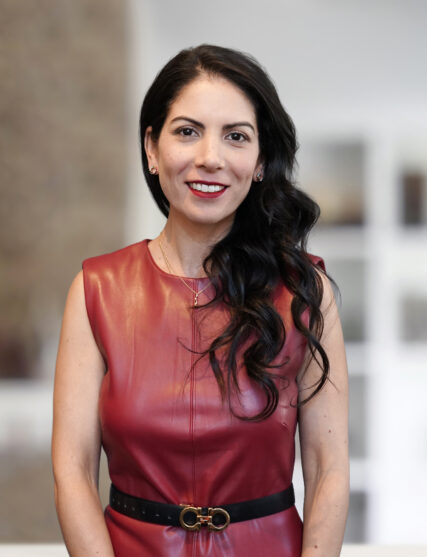 Professional portrait of a woman with long, wavy dark hair, wearing a sleeveless burgundy leather dress with a black belt, smiling against a softly blurred office interior background.