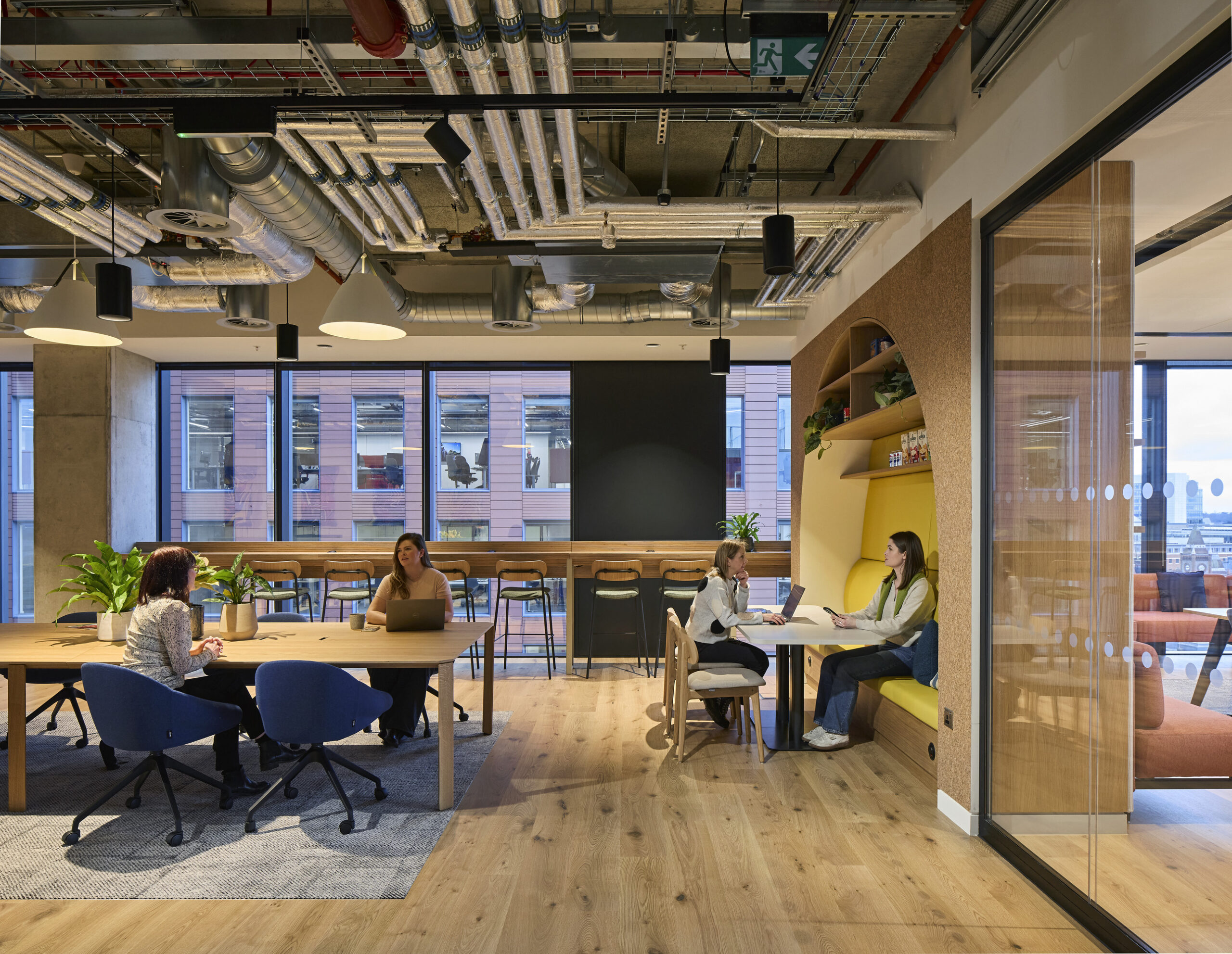 An open-plan office space features a blend of collaborative and casual seating areas. Modern wooden furniture complements the warm tones of natural light from large windows, while decorative plants enhance the inviting atmosphere. Distinctive arch-shaped shelving adds visual interest, emphasizing functional design.