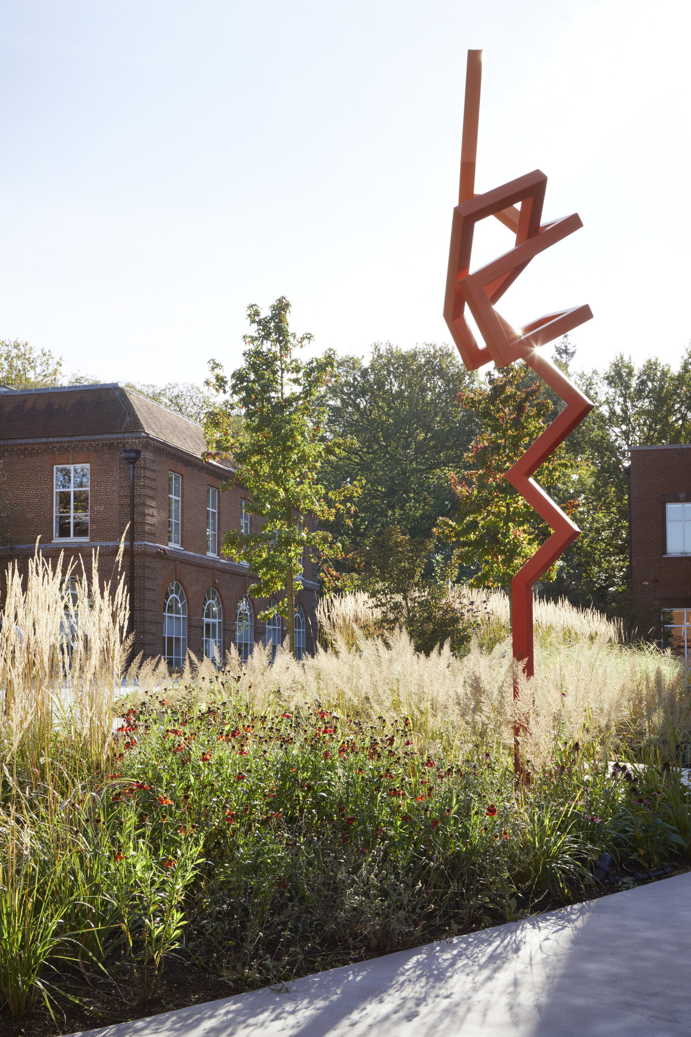 A striking red metal sculpture stands prominently in a landscaped garden, surrounded by tall grasses and vibrant flower beds. The contemporary design contrasts with the historical architecture of the adjacent building, creating a dynamic dialogue between modern art and traditional structures, enhancing the outdoor space's aesthetic appeal.