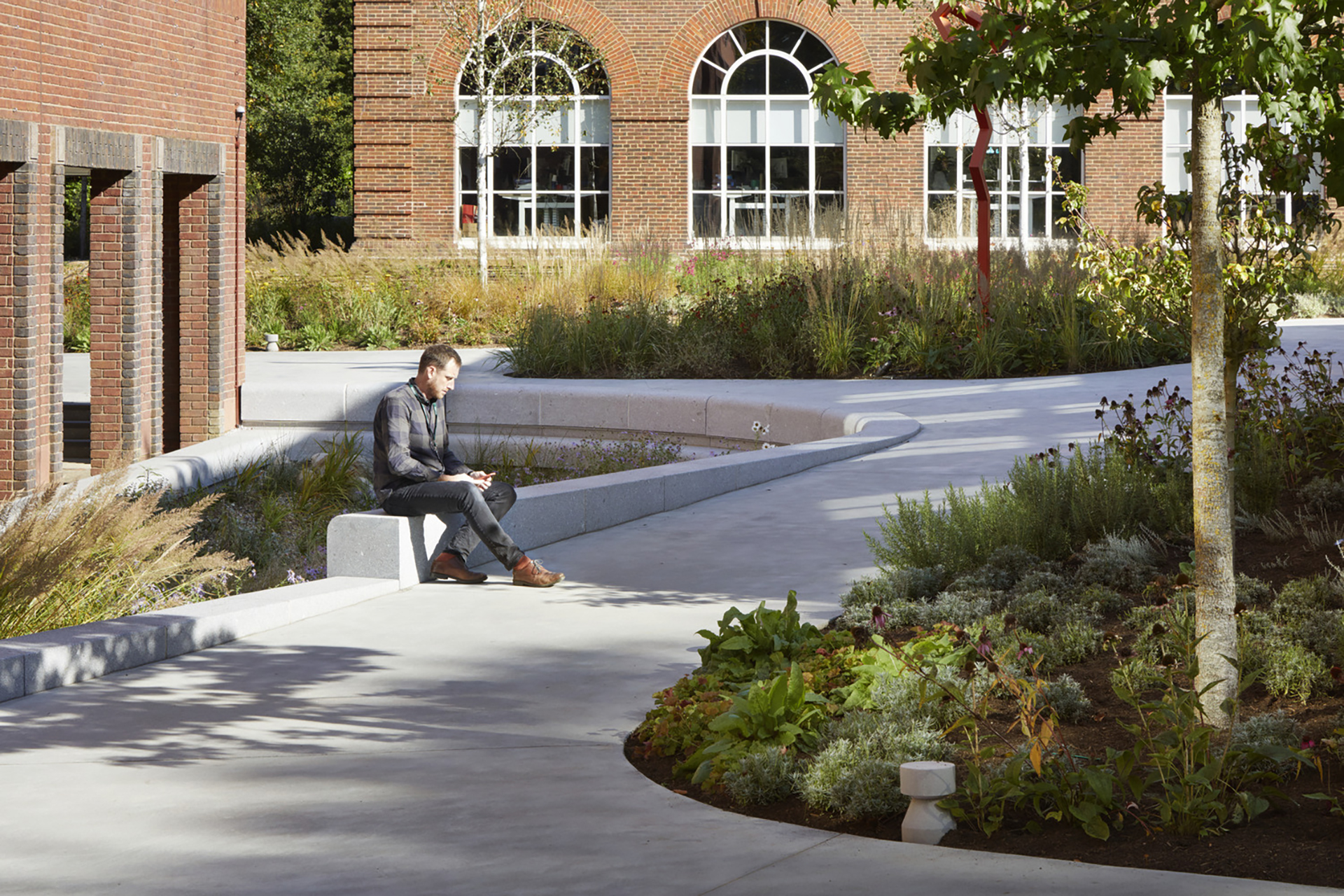 A tranquil outdoor space designed with smooth, curvilinear pathways and modern seating arrangements. A man sits on a sleek concrete bench, surrounded by meticulously arranged flora. The backdrop features large arched windows of a brick building, enhancing the inviting atmosphere of this landscaped area.