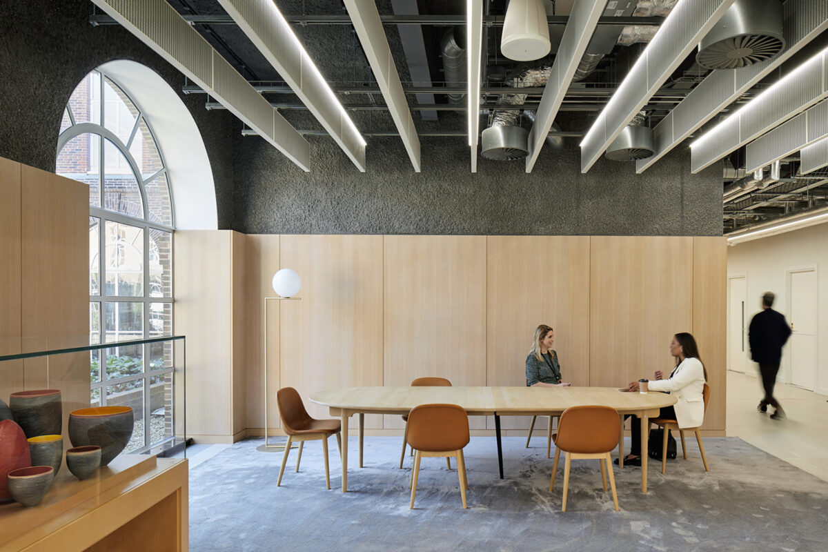 A modern meeting space featuring a large, oval wooden table surrounded by sleek, upholstered chairs. The interior showcases a warm color palette with natural wood paneling and soft textures. Large windows allow ample natural light to enhance the inviting atmosphere, facilitating collaboration among users.
