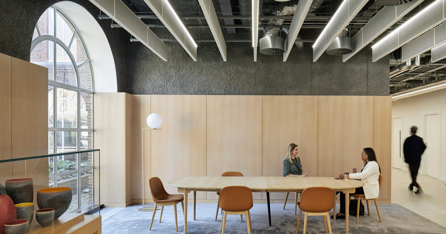 A modern meeting space featuring a large, oval wooden table surrounded by sleek, upholstered chairs. The interior showcases a warm color palette with natural wood paneling and soft textures. Large windows allow ample natural light to enhance the inviting atmosphere, facilitating collaboration among users.