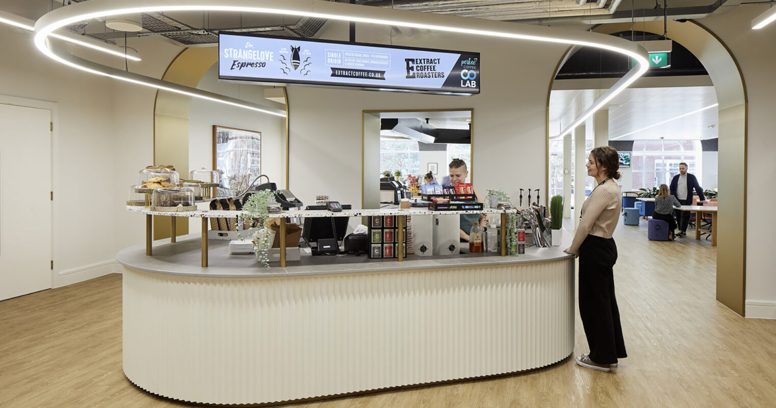 A modern coffee bar features a curved, minimalist counter with a sleek, ribbed design. Soft LED lighting highlights the space, creating a warm atmosphere. Behind the counter, various coffee and snack options are elegantly displayed, serving as a focal point in a contemporary office environment.