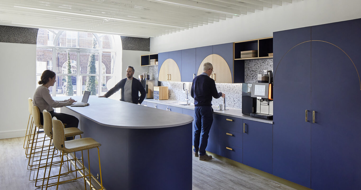 A modern kitchen featuring a curved blue bar with high gold stools. Two individuals engage at the bar, while a man prepares coffee at the cabinetry. The design includes wood accents and mosaic backsplash, complemented by natural light streaming through an arched window, creating an inviting communal space.