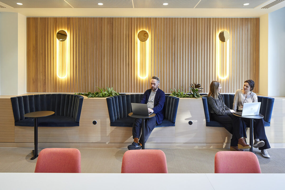 Modern workspace featuring sleek upholstered benches in rich navy, complemented by a backdrop of warm wooden paneling. Soft, atmospheric wall lights enhance the ambiance. Two professionals engage in conversation on the left, while another focuses on a laptop, showcasing a collaborative and comfortable environment designed for productivity.