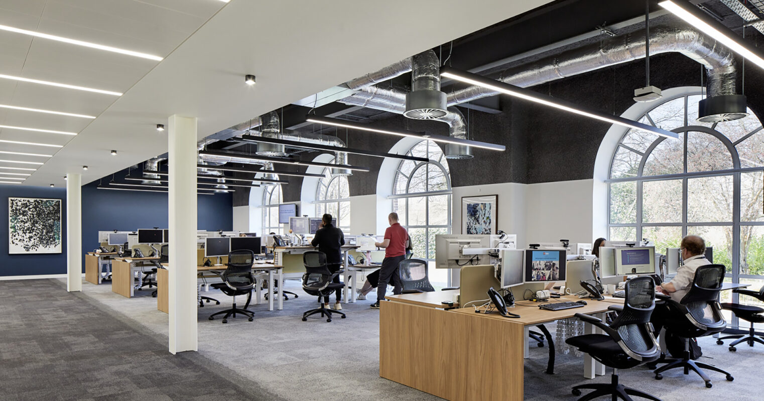 Open-plan office design featuring modern ergonomic workstations with adjustable chairs. Walls highlight contrasting colors, while large arched windows flood the space with natural light. Exposed ductwork adds an industrial touch, enhancing the contemporary feel. Collaborative areas encourage teamwork, reflecting a balance between functionality and aesthetic appeal.