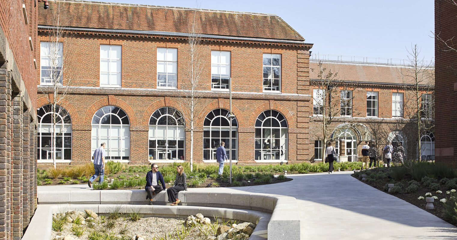A landscaped courtyard features smooth stone pathways and a central garden area with low-maintenance plants and decorative pebbles. The surrounding brick buildings showcase large arched windows, creating a harmonious blend of natural light and outdoor space, promoting relaxation and social interaction among visitors.