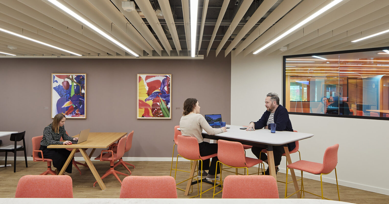 A modern collaborative workspace featuring an open layout. Light wood floors contrast with pink and grey furniture. Dynamic artwork adorns the walls, complemented by vibrant ceiling lights. Groups of professionals engage at a high-top table and traditional desks, fostering creativity and teamwork in a bright, inviting environment.