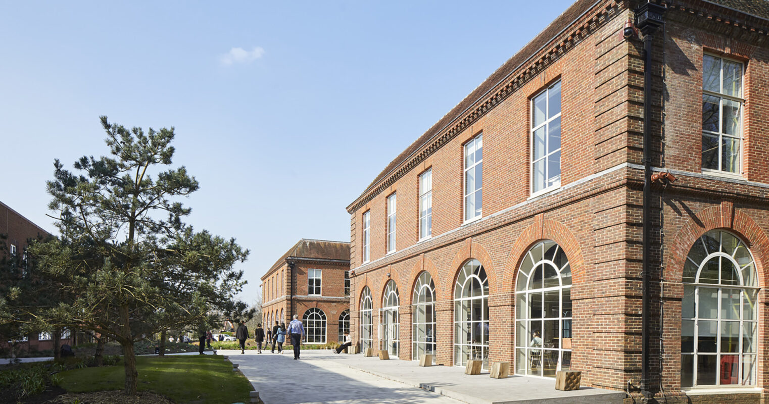 Red brick building with large arched windows, showcasing a blend of classic and contemporary architecture. A wide, paved pathway leads to the entrance, flanked by landscaped greenery and modern stone sculptures. The design emphasizes openness and integrates natural surroundings, enhancing the inviting atmosphere of the space.