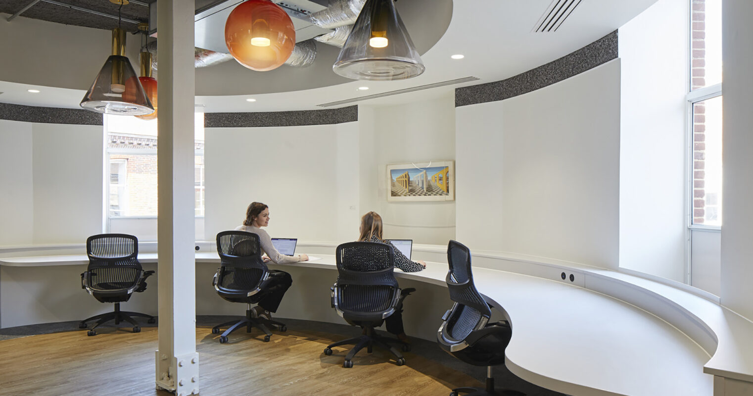 A modern workspace featuring two professionals engaged in focused work at a sleek, curved white desk. The room showcases large windows allowing natural light, accentuated by colorful pendant lights and a subtle mixture of textures in the walls, creating an inviting, collaborative atmosphere.
