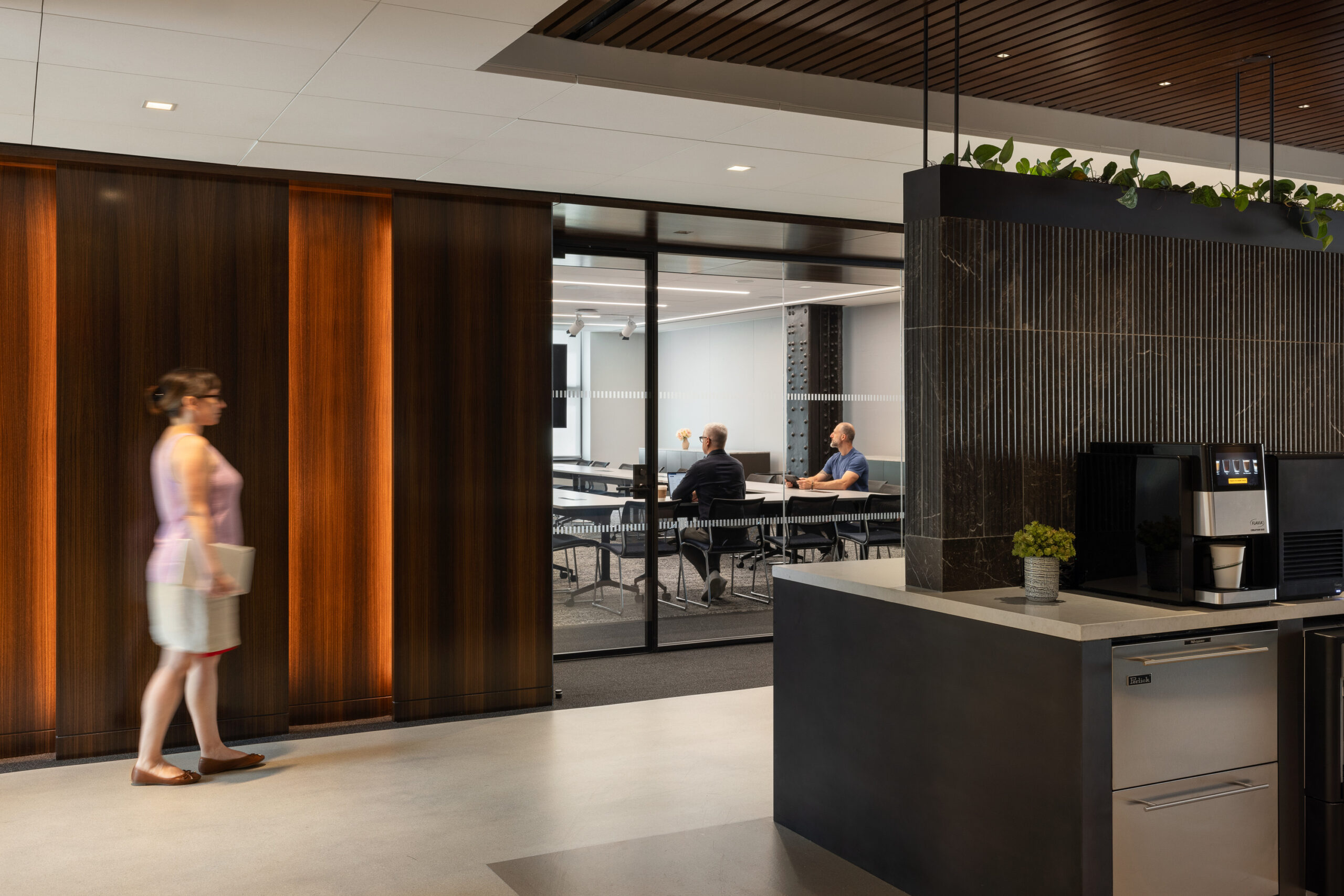 Brightly lit office interior featuring a sleek kitchen area with a stone countertop and integrated appliances. Textured wooden panels create a warm contrast with the glass-enclosed meeting room, where individuals engage in discussion. Greenery adds a natural touch, enhancing the professional yet inviting atmosphere of the space.