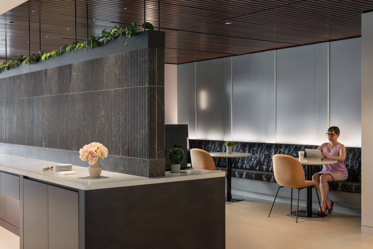 In a sophisticated reception area, warm tones and sleek materials create a welcoming ambiance. A curved, upholstered banquette pairs with modern dining chairs and a marble-topped table. Natural light enhances the space, while a textured feature wall and greenery add depth, perfect for both work and relaxation.