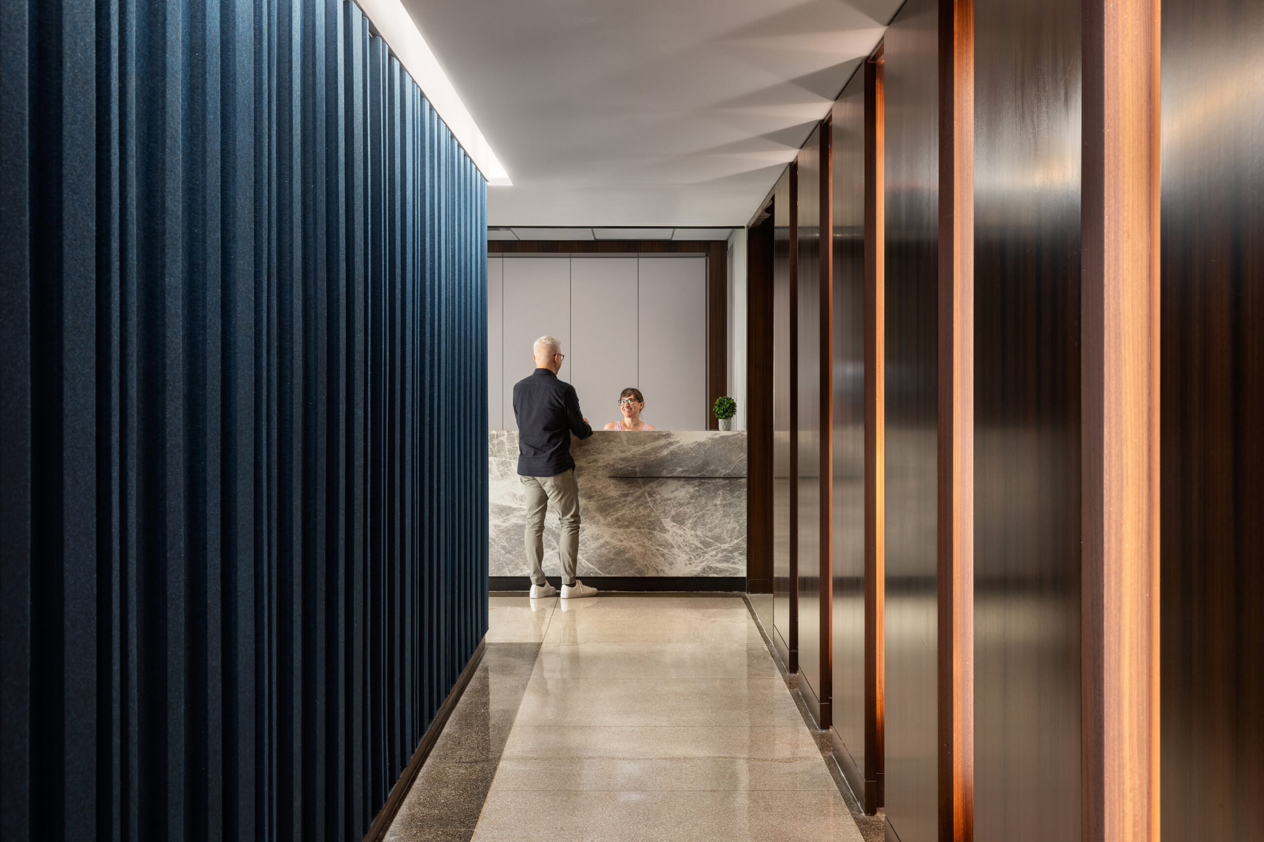 A sleek reception area features a marble front desk, complemented by warm wooden paneling and deep blue fabric accents. Soft, ambient lighting highlights the clean lines and modern architectural elements, creating an inviting atmosphere for visitors. The corridor's design efficiently guides guests towards the front desk.