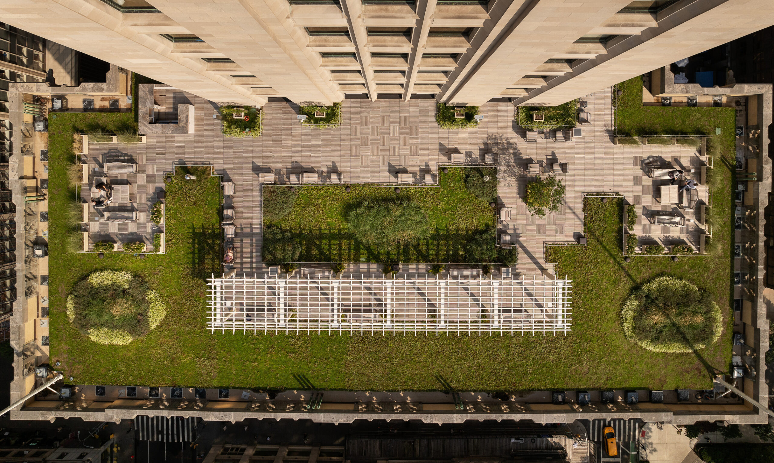 Aerial view of a city rooftop garden, featuring an expansive green space with meticulously arranged plant beds and seating areas. The layout includes pathways, decorative trellises, and sections of grass, creating a tranquil retreat amid urban architecture. Natural elements enhance the overall aesthetic and promote sustainability.