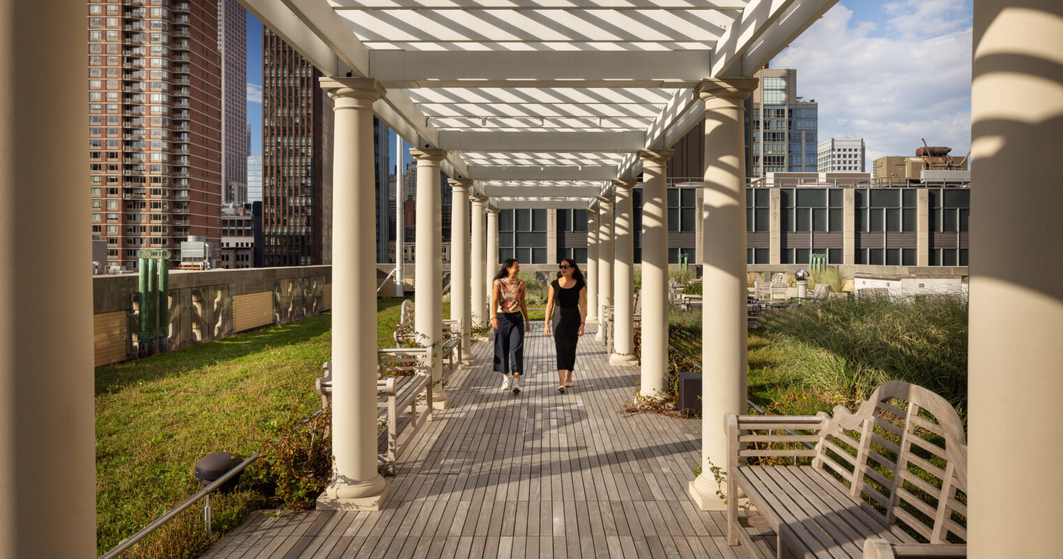 A stylish urban rooftop garden features a covered walkway supported by elegant columns, casting intricate shadows on the wooden decking. Lush greenery flanks the pathway, while two individuals stroll leisurely, enjoying the serene atmosphere amid the cityscape backdrop and modern architecture.