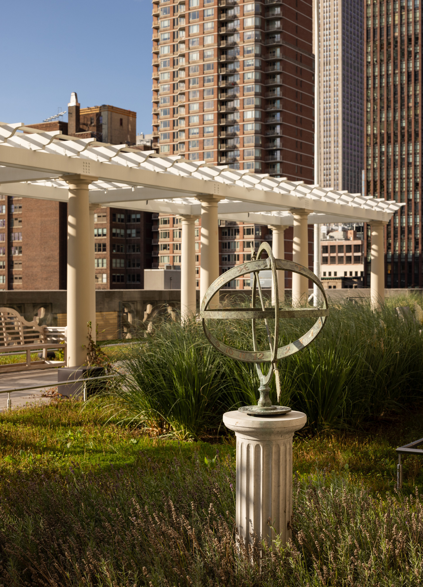 A contemporary rooftop garden showcases a sleek design with a metal sundial on a stone pedestal, surrounded by lush greenery. The pergola provides shade, enhancing the outdoor space's elegance, while urban skyscrapers form a striking backdrop, blending nature with modern city living.