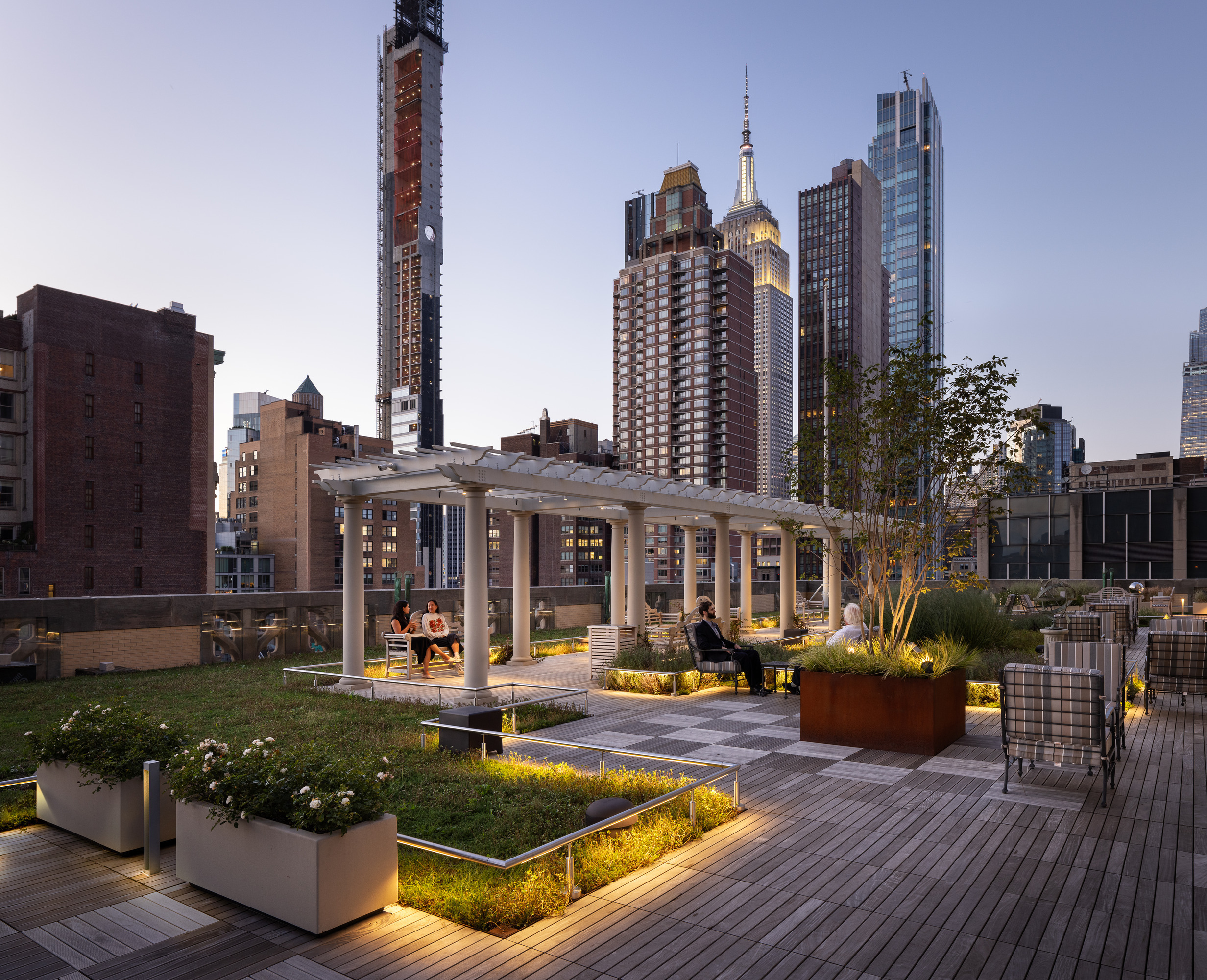 A contemporary rooftop garden features sleek wooden decking, integrated seating, and lush planters, offering a serene urban oasis. Backlit by subtle landscape lighting, the design showcases a harmonious blend of greenery and city skyline, highlighting modern architecture, relaxation zones, and stunning evening views of skyscrapers.