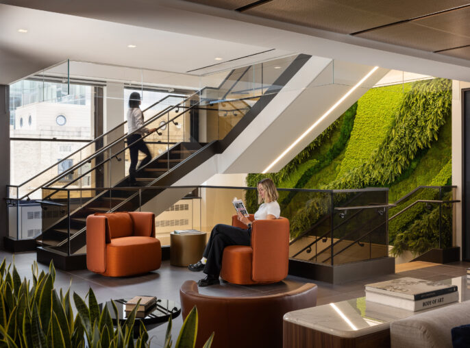 Contemporary lobby featuring curved orange seating and natural greenery accentuating a vertical wall. A woman enjoys reading while seated next to a round wooden table, framed by glass railings and modern staircase design. The space blends functionality with inviting aesthetics, encouraging relaxation and collaboration.