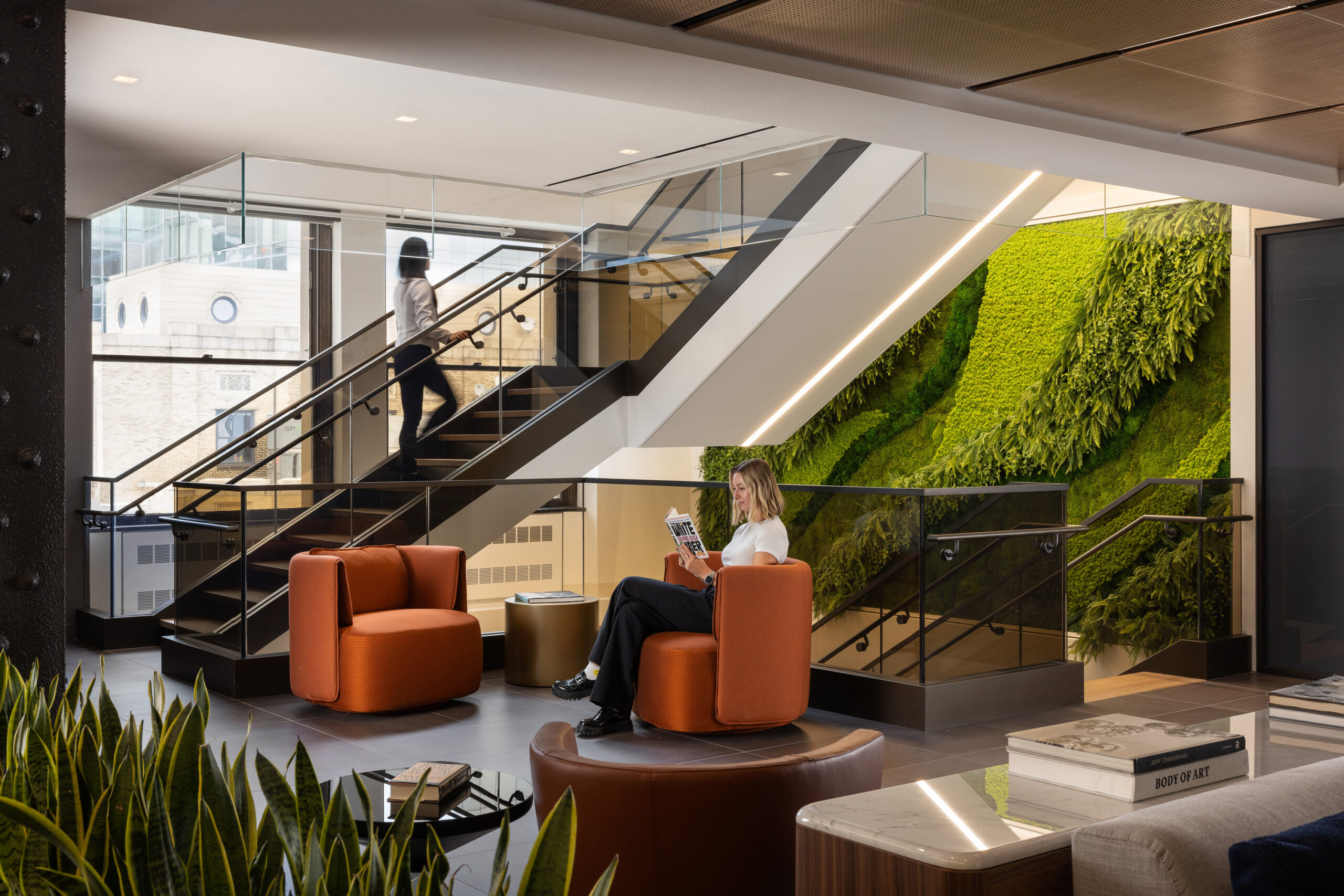 Contemporary lobby featuring curved orange seating and natural greenery accentuating a vertical wall. A woman enjoys reading while seated next to a round wooden table, framed by glass railings and modern staircase design. The space blends functionality with inviting aesthetics, encouraging relaxation and collaboration.