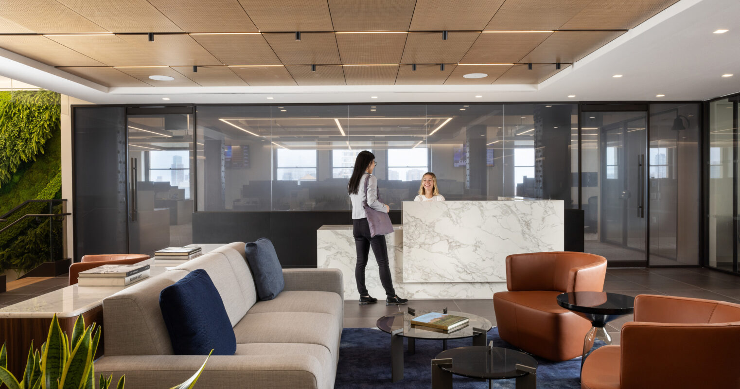 A modern office reception area features a large white marble reception desk, complemented by warm wood paneling on the ceiling. Stylish orange chairs and a light grey sofa offer comfort, while a vertical green wall brings a touch of nature, enhancing the inviting atmosphere for clients and visitors.