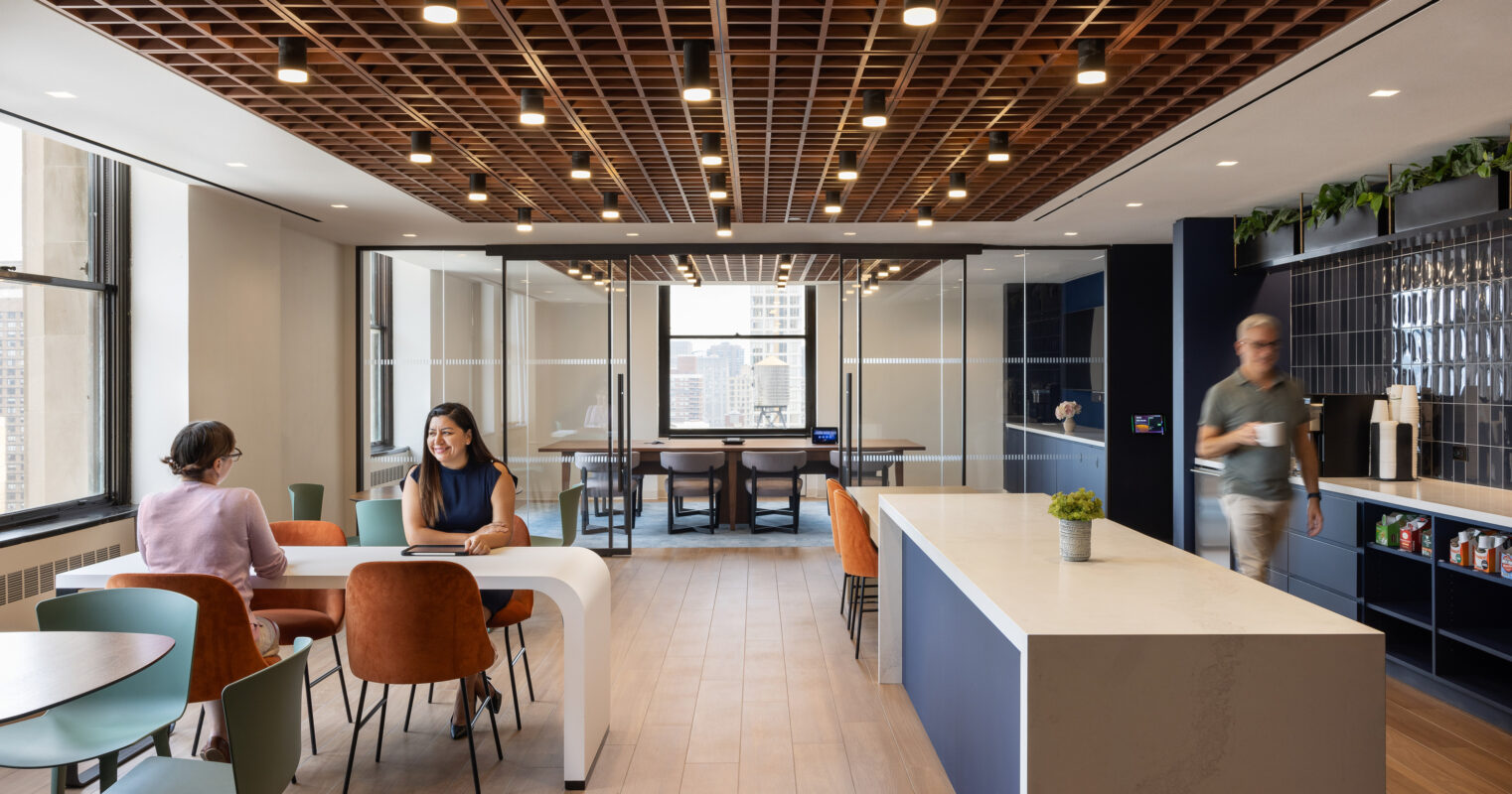 A spacious modern office showcasing an open-layout design. Natural light floods through large windows, illuminating sleek furnishings. Collaborative areas feature round tables and colorful chairs, while a central kitchen counter offers refreshments. A geometric wood ceiling adds texture and warmth to the contemporary aesthetic.